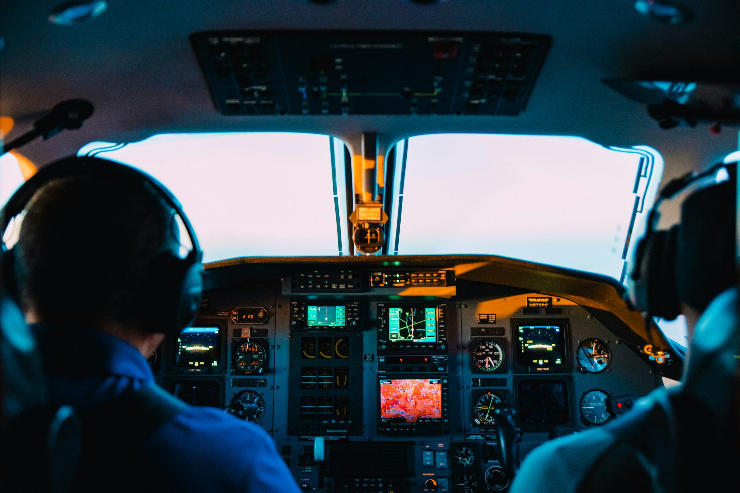 The image shows the interior of an airplane cockpit with two pilots seated. They are wearing headsets and facing a panel filled with various instruments, gauges, and screens. The view through the cockpit windows shows a bright sky, suggesting the plane is in flight.