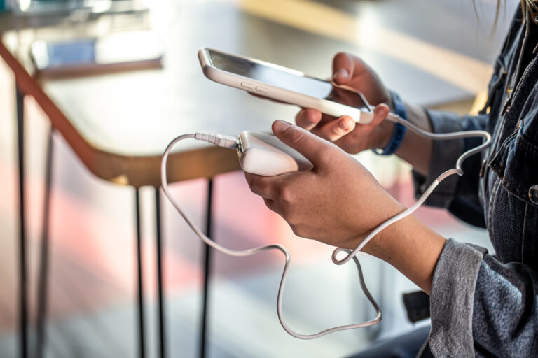 A person is holding a smartphone and a white computer mouse, with a cable connecting the two. The person is wearing a denim jacket, and the background shows a blurred table and chair.