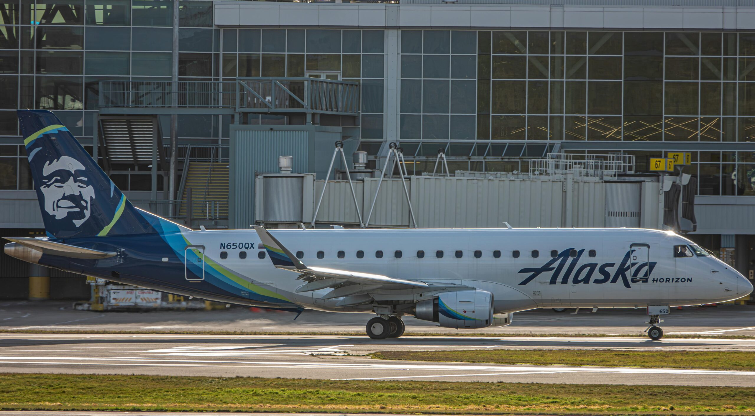 The image shows an Alaska Horizon airplane on the tarmac in front of an airport terminal. The aircraft is painted in the airline's livery, featuring the word "Alaska" on the fuselage and a stylized face on the tail. The terminal building in the background has large windows and various airport equipment is visible.