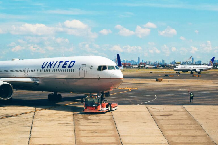 The image shows a United Airlines airplane on the tarmac at an airport. The aircraft is being towed by a red vehicle. In the background, another United Airlines plane is visible, along with a city skyline under a blue sky with scattered clouds. A person in a safety vest is standing on the tarmac, holding a signaling wand.