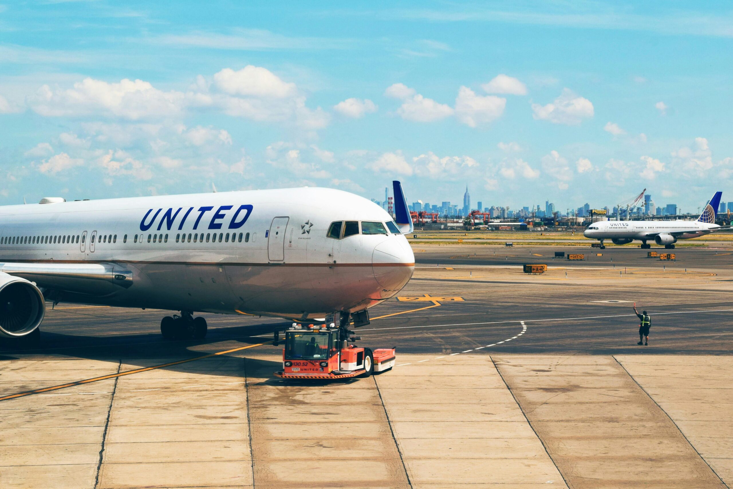 The image shows a United Airlines airplane on the tarmac at an airport. The aircraft is being towed by a red vehicle. In the background, another United Airlines plane is visible, along with a city skyline under a blue sky with scattered clouds. A person in a safety vest is standing on the tarmac, holding a signaling wand.