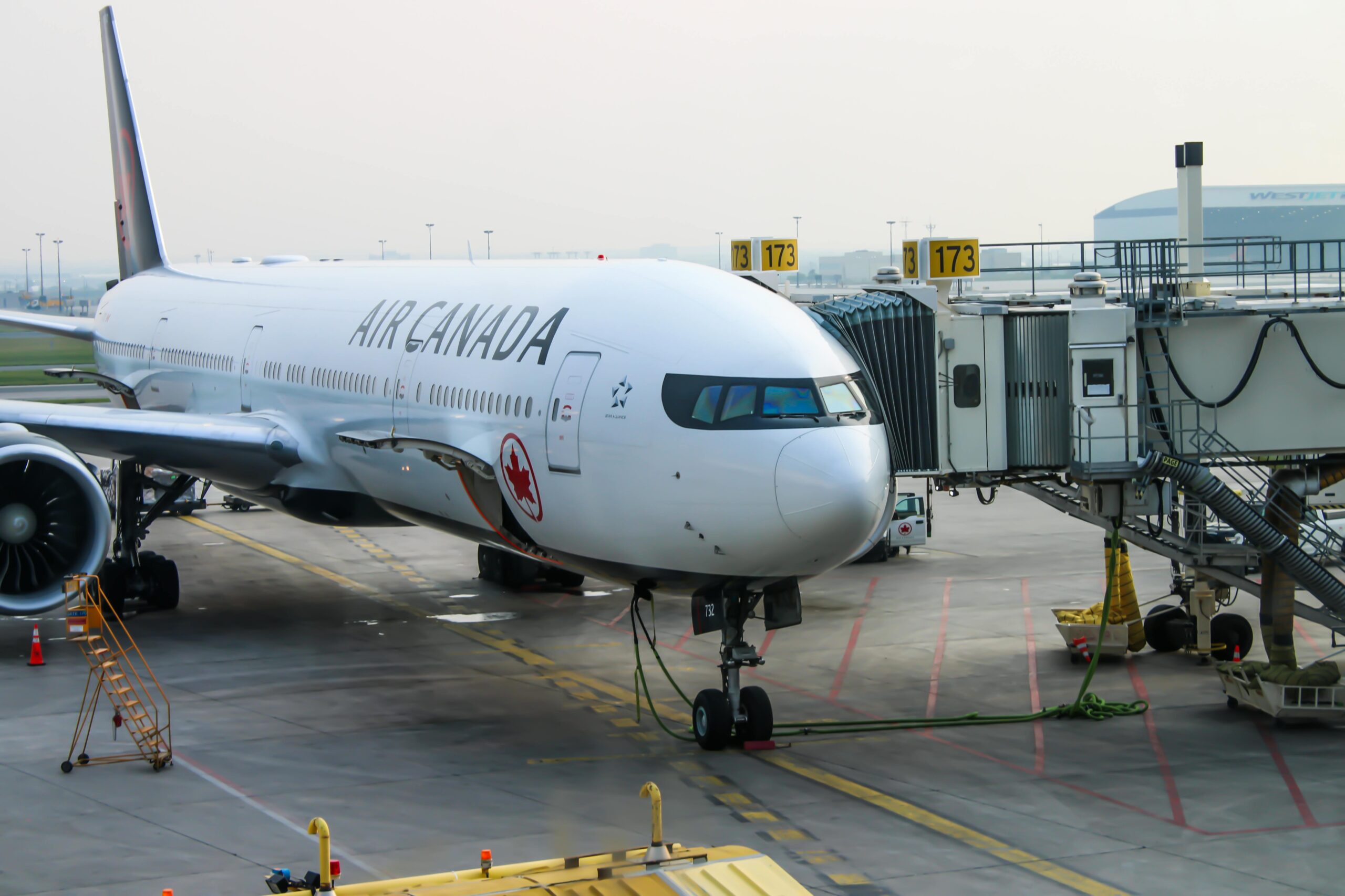 An Air Canada airplane is parked at an airport gate, connected to a jet bridge. The aircraft is a large passenger jet with visible engines and landing gear. The airport tarmac and terminal are in the background, with some signage and equipment nearby.