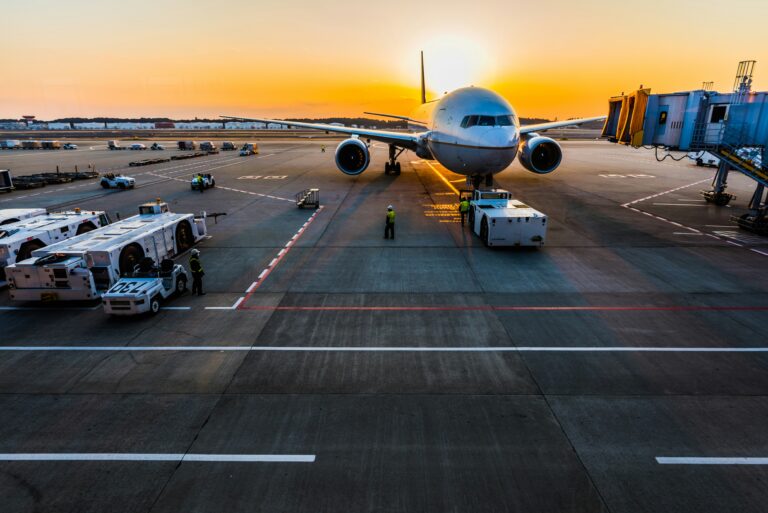 A large airplane is parked on an airport tarmac at sunset. The sun is setting behind the aircraft, casting a warm glow. Ground crew members in high-visibility vests are working around the plane, and various airport vehicles are nearby. A jet bridge is connected to the aircraft on the right side.