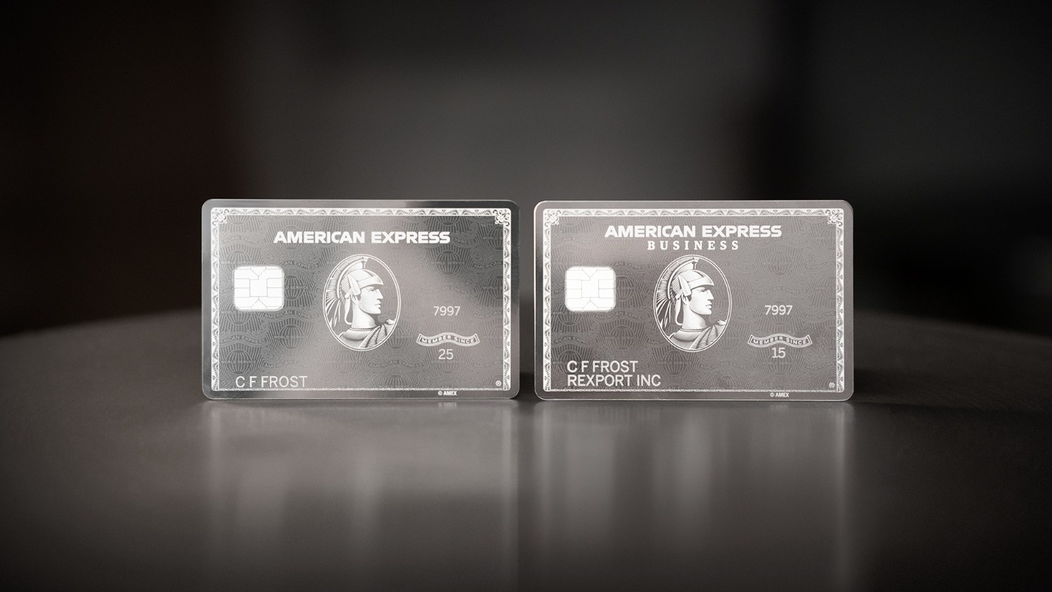 The image shows two American Express credit cards placed side by side on a dark surface. The card on the left is a personal card, and the one on the right is a business card. Both cards have a silver design with the American Express logo and a depiction of a centurion. The cards have chip technology and display the name "C F Frost."