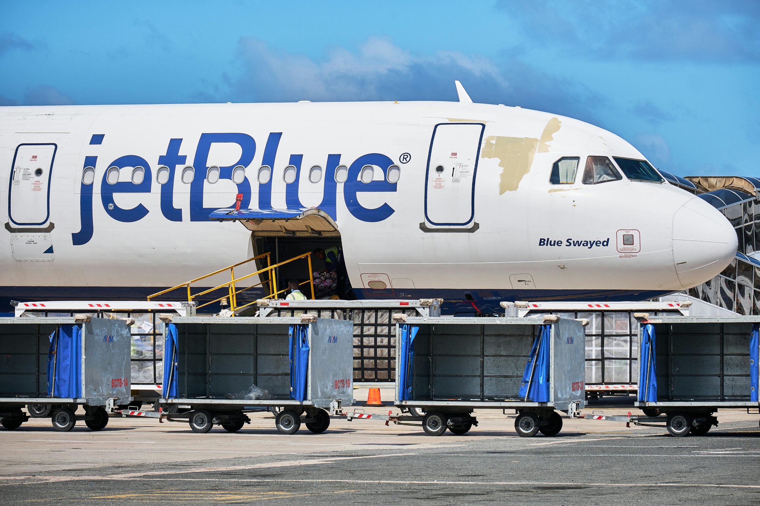 A JetBlue airplane is parked on the tarmac with its cargo door open. Several baggage carts are lined up in front of the aircraft. The sky is clear and blue in the background.