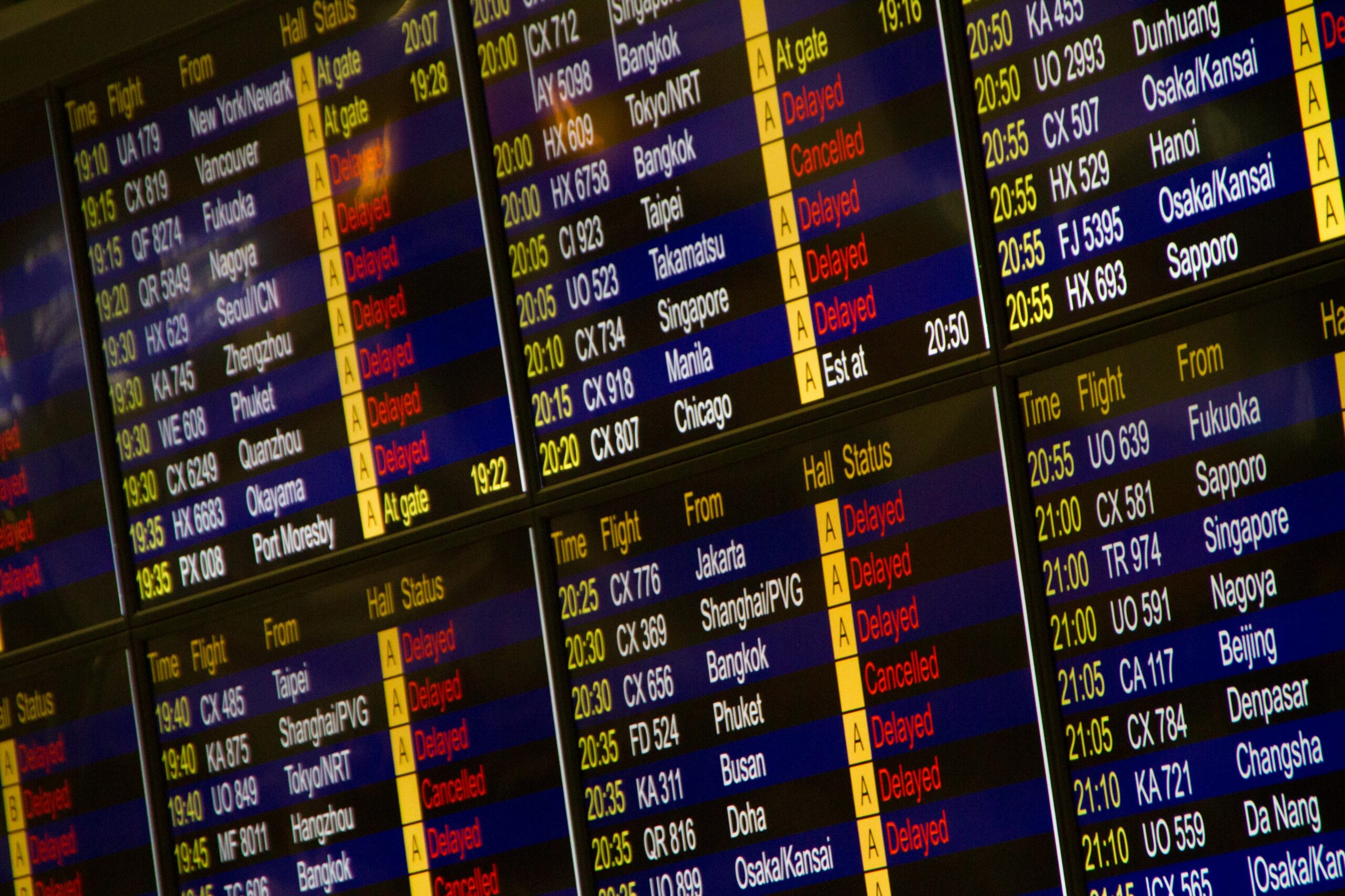 The image shows an airport departure board displaying flight information. It includes columns for time, flight number, origin, hall, and status. Many flights are marked as "Delayed" in red, with one marked as "Cancelled" in red, and a few listed as "At gate" in yellow. The board shows flights from various international locations such as New York, Vancouver, Fukuoka, and others.