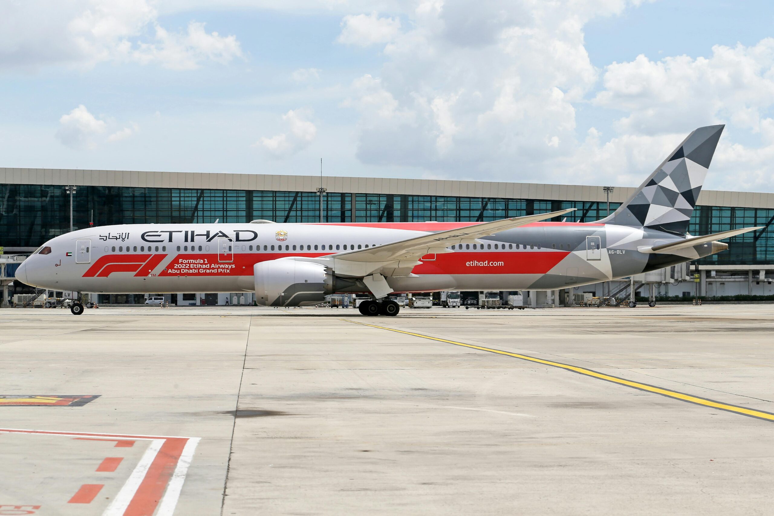 A passenger airplane with Etihad Airways branding is parked on an airport tarmac. The aircraft features a special livery promoting the "Formula 1 2022 Etihad Airways Abu Dhabi Grand Prix," with a red and white color scheme and geometric patterns on the tail. The background shows a modern airport terminal with large glass windows under a partly cloudy sky.