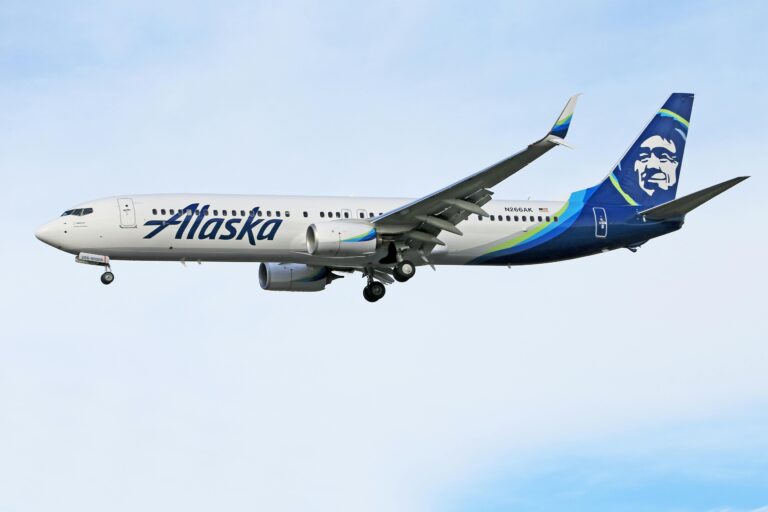 A commercial airplane in flight against a clear sky. The aircraft is white with "Alaska" written on the side in blue letters. The tail features a stylized face logo in blue and green. The landing gear is extended.
