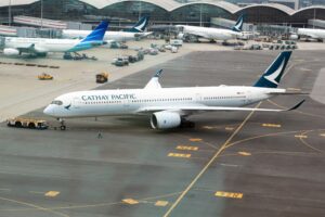The image shows a Cathay Pacific airplane on the tarmac at an airport. The aircraft is being towed by a vehicle. In the background, there are other airplanes, including one from Garuda Indonesia, parked near the terminal. The airport terminal building is visible with large glass windows. There are various ground service vehicles and equipment around the planes.