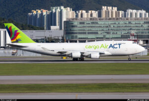 A large cargo airplane is taxiing on a runway at an airport. The aircraft is white with "cargo Air ACT" written on the side in green and blue letters. The tail features a colorful design with red, purple, and green elements on a lime green background. In the background, there are modern buildings and a mountainous landscape.