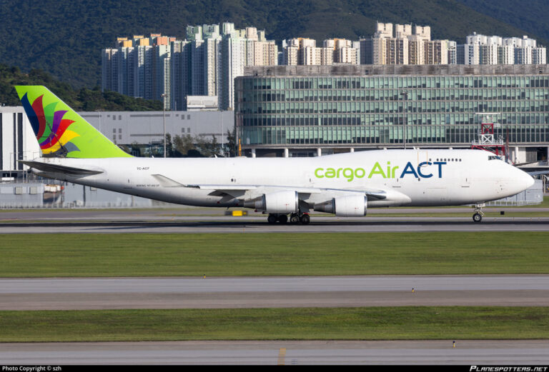 A large cargo airplane is taxiing on a runway at an airport. The aircraft is white with "cargo Air ACT" written on the side in green and blue letters. The tail features a colorful design with red, purple, and green elements on a lime green background. In the background, there are modern buildings and a mountainous landscape.