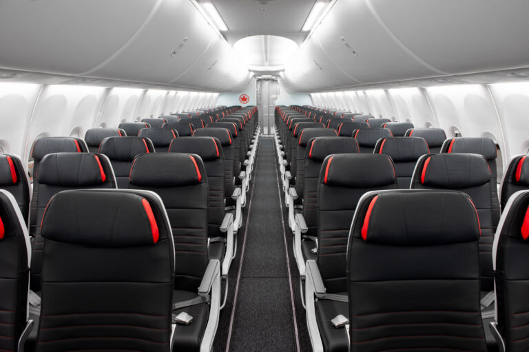 The image shows the interior of an empty airplane cabin. The seats are arranged in rows on either side of a central aisle. The seats are upholstered in black with red accents. Overhead compartments are closed, and the lighting is bright, giving a clean and modern appearance.