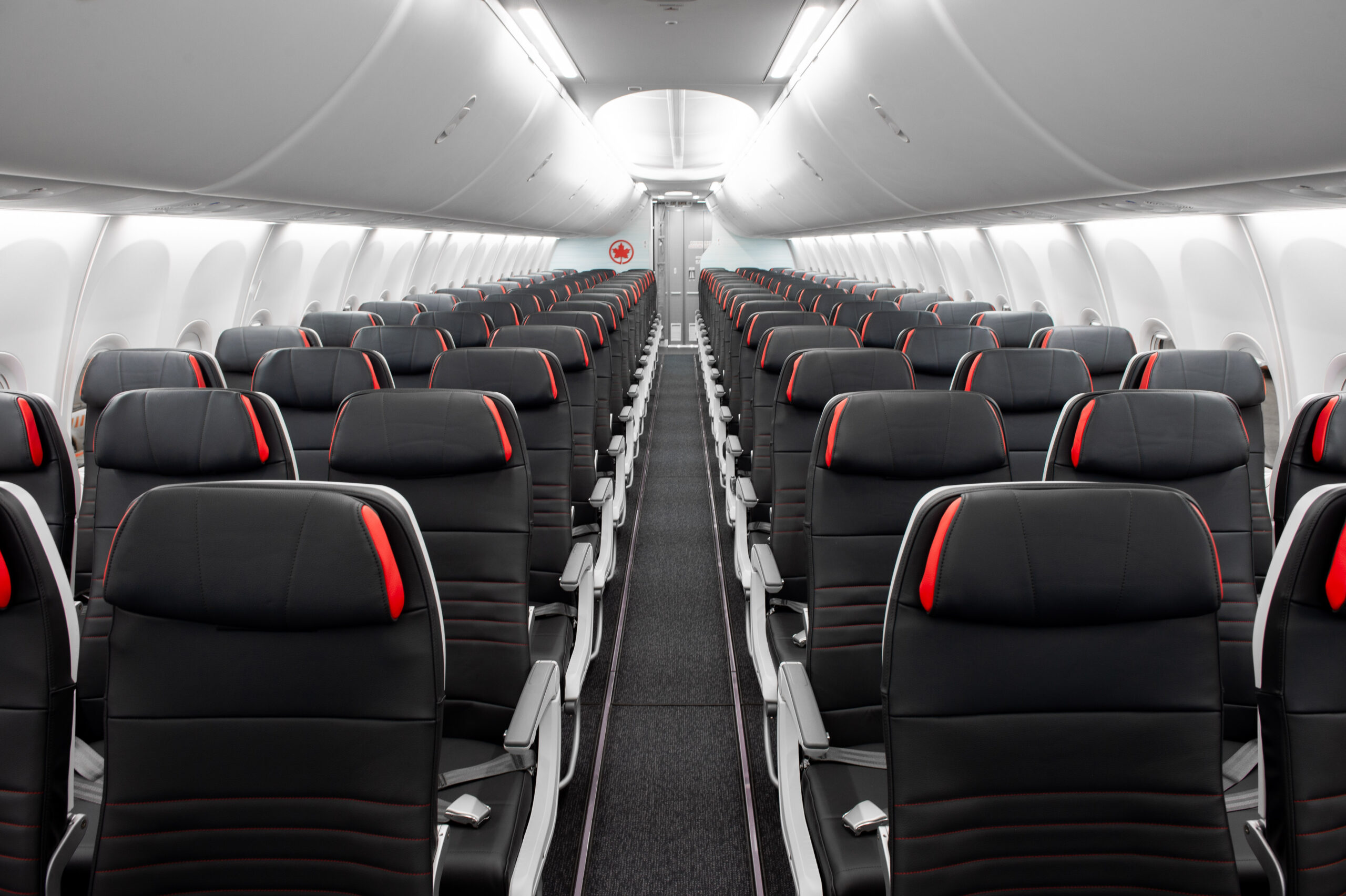 The image shows the interior of an empty airplane cabin. The seats are arranged in rows on either side of a central aisle. The seats are upholstered in black with red accents. Overhead compartments are closed, and the lighting is bright, giving a clean and modern appearance.