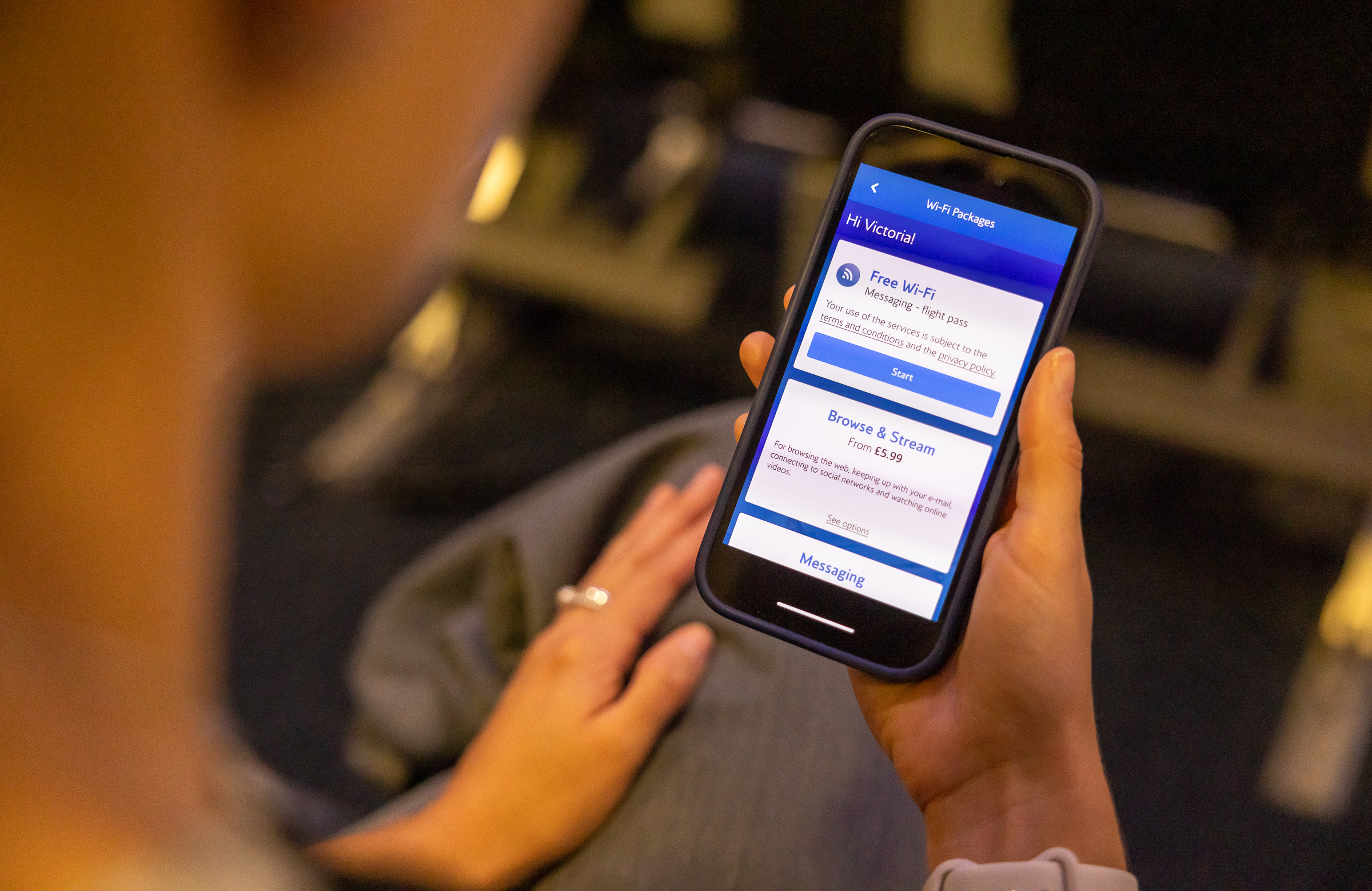 A person is holding a smartphone displaying a Wi-Fi package selection screen. The screen shows options for "Free Wi-Fi" for messaging with a flight pass and a "Browse & Stream" option starting from £5.99. The background is blurred, suggesting the person might be on an airplane.