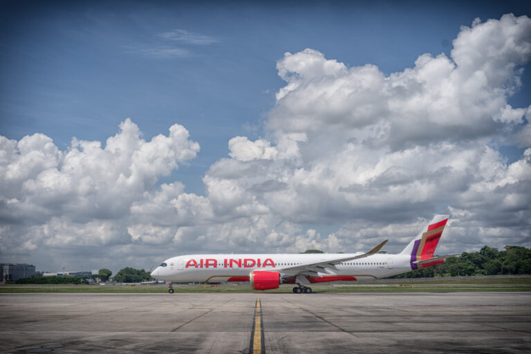 The image shows an Air India airplane on a runway. The aircraft is white with red accents, and the words "AIR INDIA" are prominently displayed on the side. The sky is blue with scattered white clouds, and there is greenery in the background.