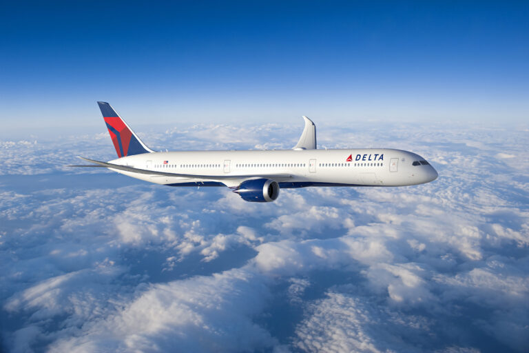 A Delta Air Lines airplane is flying above the clouds. The aircraft is white with the Delta logo on the fuselage and a red and blue tail. The sky is clear and blue.