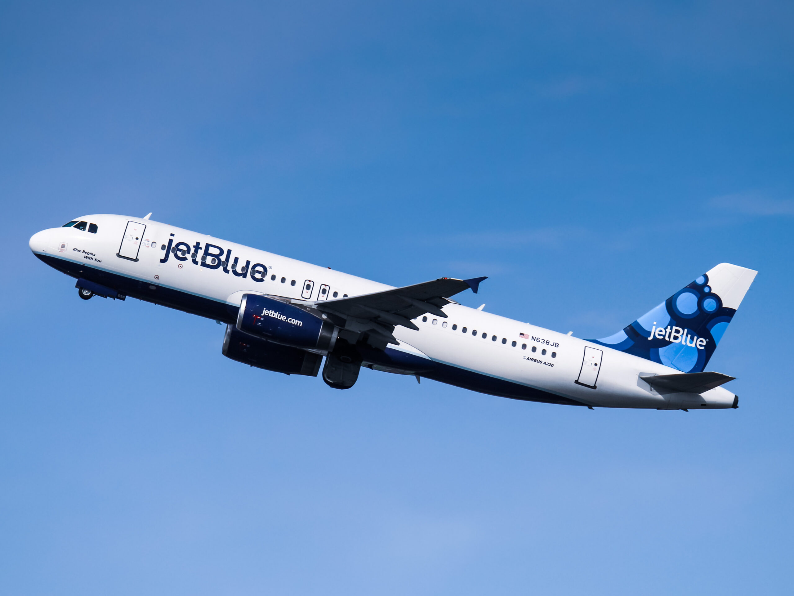 A jetBlue Airbus A320 airplane is flying in a clear blue sky. The aircraft is white with blue accents, and the jetBlue logo is visible on the fuselage and tail. The engines are also marked with the jetBlue branding.